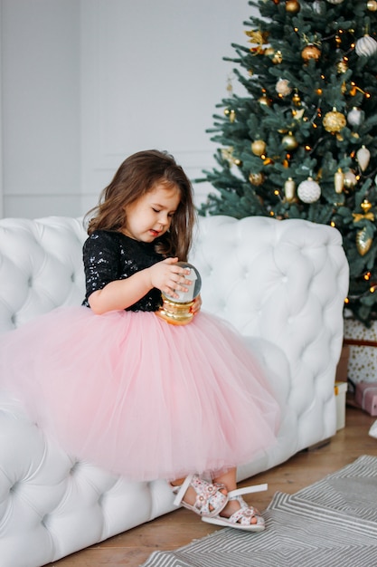 Kleines Mädchen im schönen Kleiderballettröckchenrock mit Glaskugel im Wohnzimmer an des Weihnachtsbaums