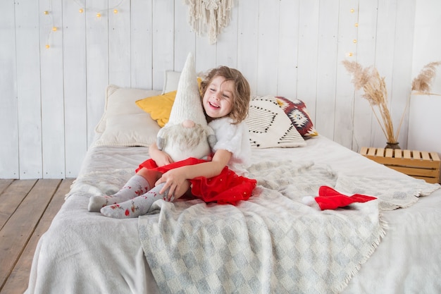 Kleines Mädchen im Weihnachtsmannkostüm mit Weihnachtsmannspielzeug, das auf dem Bett sitzt.