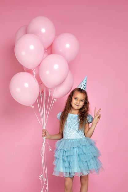 Kleines Mädchen in blauem Kleid und Geburtstagshut, das mit pastellrosa Luftballons feiert