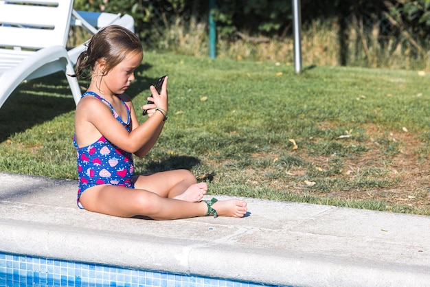 Kleines Mädchen in einem Badeanzug sitzt am Rand des Pools und schaut auf ihr Handy Kindheit Kinder Smartphone Technologie Urlaub Internet und Spaß Konzept