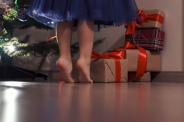 Kleines Mädchen in einem blauen Kleid steht auf Zehenspitzen am Weihnachtsbaum, die Beine hautnah
