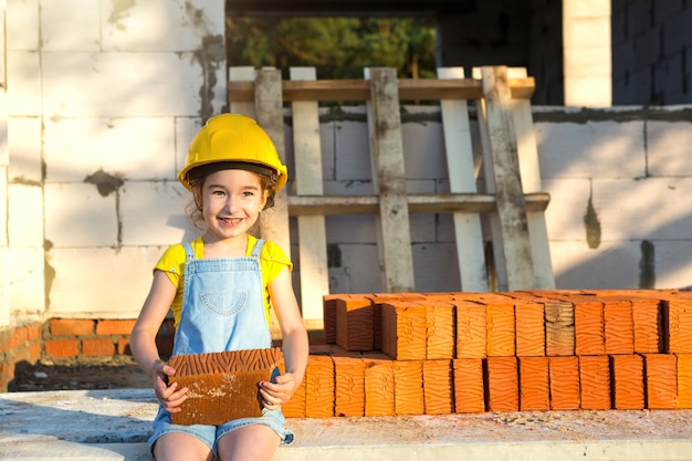 Kleines Mädchen in einem gelben Bauarbeiterhelm spielt Baumeister auf der Baustelle ihres zukünftigen Hauses. Umzugserwartung, Berufswahl, Kindererziehung. Zukünftiger Beruf, Familienunternehmen.