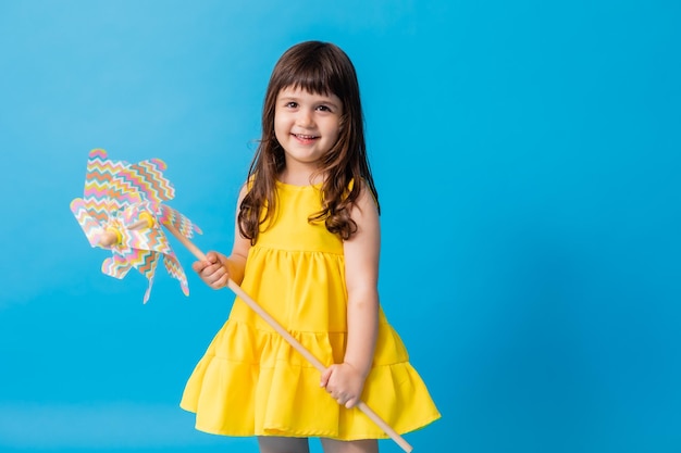 Kleines Mädchen in einem gelben Kleid auf blauem Hintergrund hält eine Spielzeugwindmühle für Kinder in ihren Händen