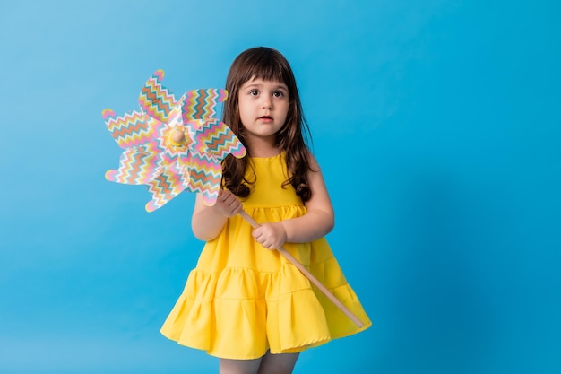 Kleines Mädchen in einem gelben Kleid auf blauem Hintergrund hält eine Spielzeugwindmühle für Kinder in ihren Händen