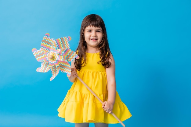 Kleines Mädchen in einem gelben Kleid auf blauem Hintergrund hält eine Spielzeugwindmühle für Kinder in ihren Händen