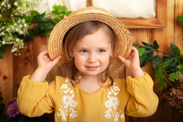 Kleines Mädchen in einem gelben Kleid lächelt und hält einen Strohhut. Kindheitskonzept. Porträt des niedlichen kleinen blonden Mädchens in der Landschaft, Sommerzeit. Schönes Kind im Frühlingsgarten. Rustikal. Gartenarbeit