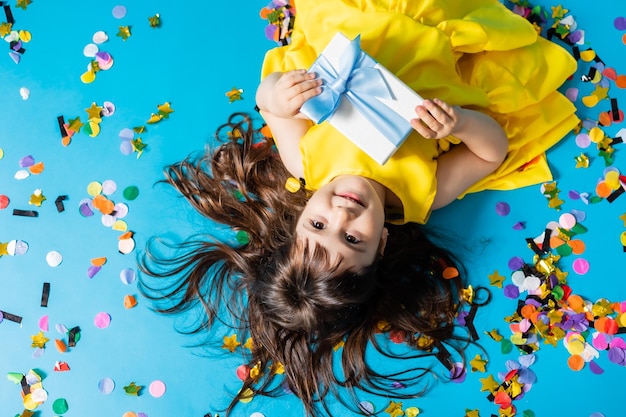 Kleines Mädchen in einem gelben Kleid liegt auf dem Boden mit einer blauen Geschenkbox in ihren Händen Geburtstag