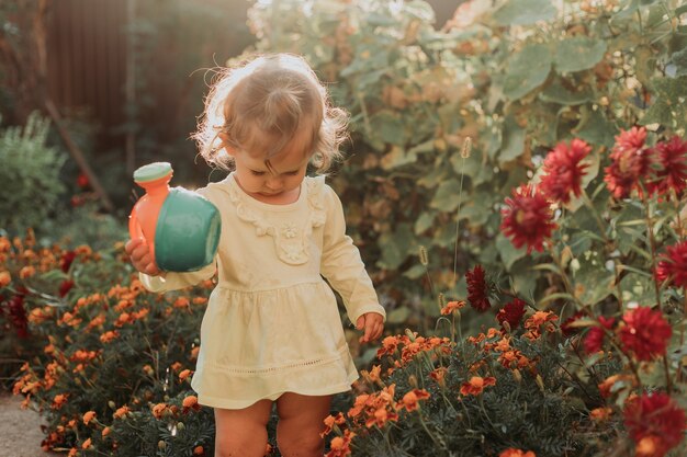 Kleines Mädchen in einem gelben Kleid und Gummistiefeln gießt Blumen im Garten Kleiner Helfer