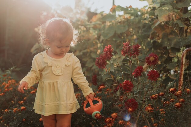 Kleines Mädchen in einem gelben Kleid und Gummistiefeln gießt Blumen im Garten Kleiner Helfer