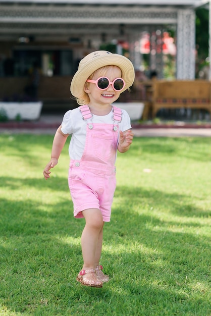 Kleines Mädchen in einem Hut und in der rosa Sonnenbrille, die Spaß haben und auf dem grünen Gras spielen. Sorgloses Kindheitskonzept.