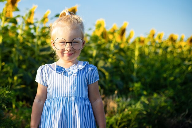 Kleines Mädchen in einem Kleid und in den Gläsern auf dem Hintergrund eines Sonnenblumenfeldes.