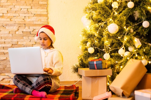 Kleines Mädchen in einem Pullover für das neue Jahr. Ein Mädchen und ein Laptop am Weihnachtsbaum mit einem Buch zu Weihnachten. Ein Kind und ein Computer für das neue Jahr.