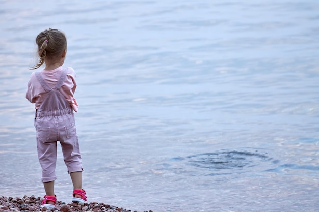 Kleines Mädchen in einem rosafarbenen Overall steht allein am Strand und blickt in die Ferne