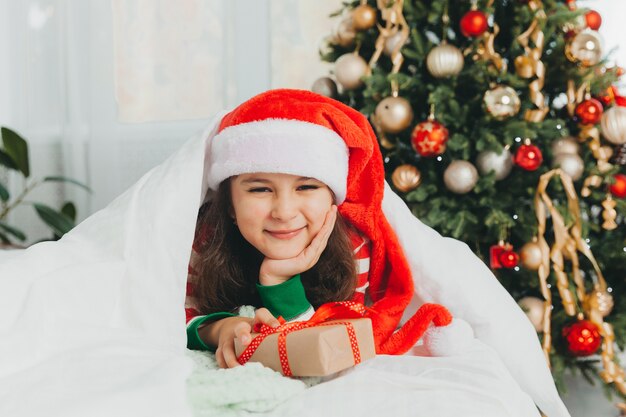 Kleines Mädchen in einem roten Hut des neuen Jahres mit einem Weihnachtsgeschenk. Sie liegt auf dem Bett und umarmt eine Schachtel vor dem Hintergrund eines Weihnachtsbaums.