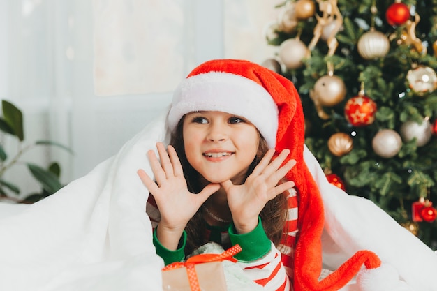 Kleines Mädchen in einem roten Hut des neuen Jahres mit einem Weihnachtsgeschenk. Sie liegt auf dem Bett und umarmt eine Schachtel vor dem Hintergrund eines Weihnachtsbaums.