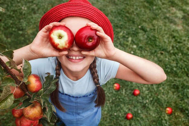 Kleines Mädchen in einem roten Hut hält zwei Äpfel in der Nähe der Augen auf einem Grashintergrund