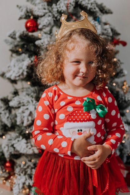 Kleines Mädchen in einem roten Karnevalskleid und einer Krone spielt mit Spielzeughirschen neben dem Weihnachtsbaum