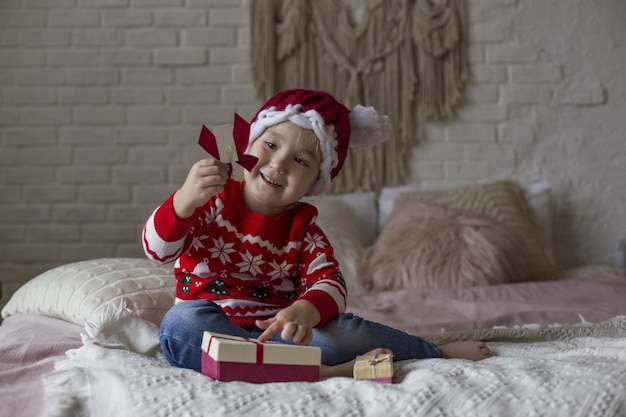 Kleines Mädchen in einem roten roten Weihnachtspullover und in der Weihnachtsmannmütze spielt auf dem Bett mit Geschenken