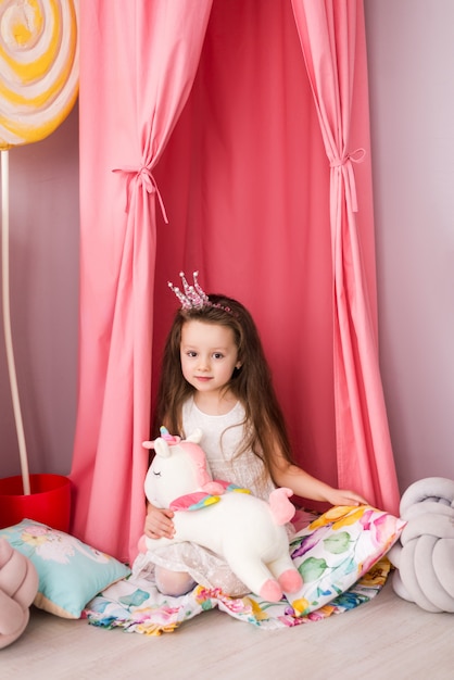 kleines Mädchen in einem schicken Kleid auf dem Hintergrund eines schönen Kinderzimmers. Spielzeug Einhorn in seinen Händen.