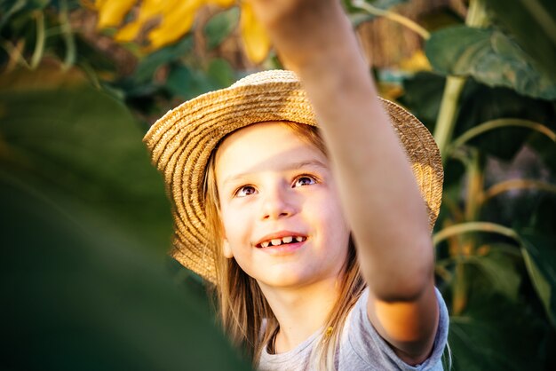 Kleines Mädchen in einem Strohhut, der in den Sonnenblumenfeldkindern unter Sonnenblumensommerhintergrund spielt