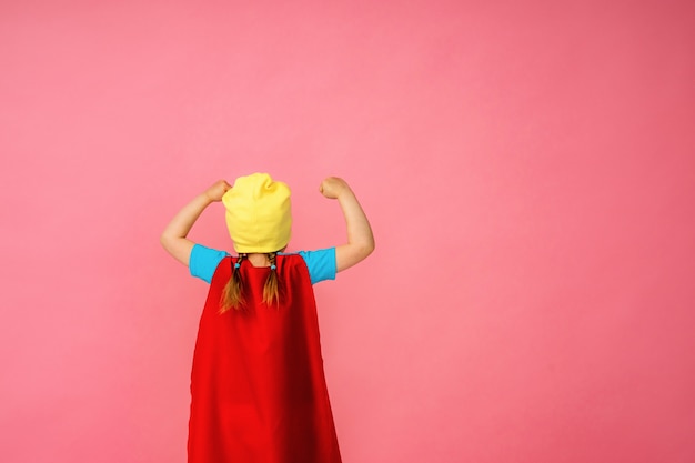 Kleines Mädchen in einem Superheldenkostüm in rotem Umhang und Hut zeigt, wie stark sie ist.