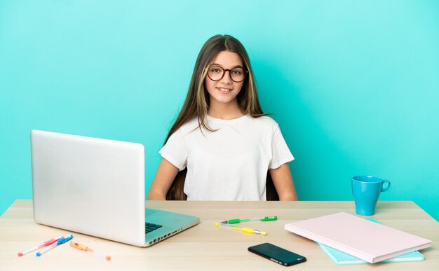 Kleines Mädchen in einem Tisch mit einem Laptop über isoliertem blauem Hintergrund lachen