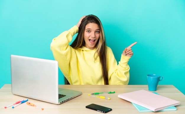 Kleines Mädchen in einem Tisch mit einem Laptop über isoliertem blauem Hintergrund überrascht und zeigt mit dem Finger zur Seite