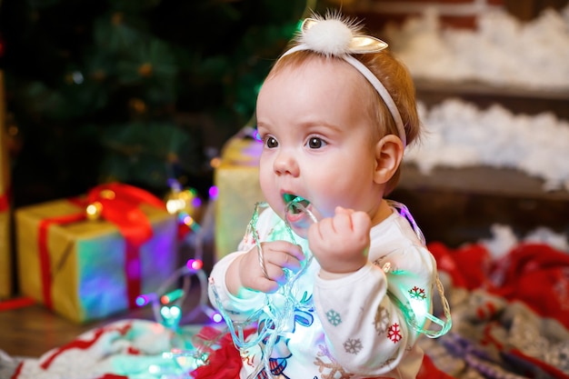 Kleines Mädchen in einem warmen Pullover sitzt unter einem Weihnachtsbaum mit Spielzeug und Geschenken. Glückliche Kindheit. Silvester festliche Atmosphäre