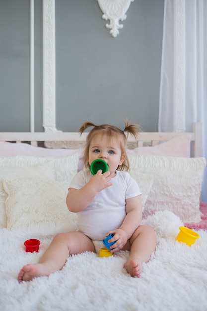 Kleines Mädchen in einem weißen Body spielt auf dem Bett mit bunten Spielzeugen