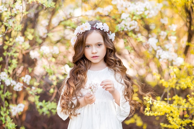 Kleines Mädchen in einem weißen Kleid auf einem Hintergrund der gelben Blumen. Frühlingsfoto mit einem schönen Mädchen. Ein Kind mit einem Blumenkranz ..