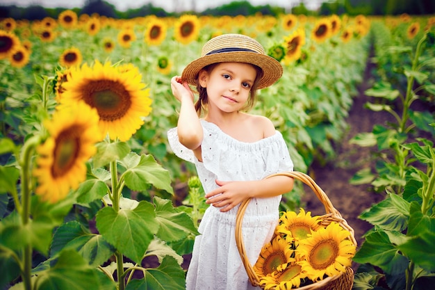 Kleines Mädchen in einem weißen Kleid, ein Strohhut mit einem Korb voller Sonnenblumen, die in die Kamera in einem Feld lächeln