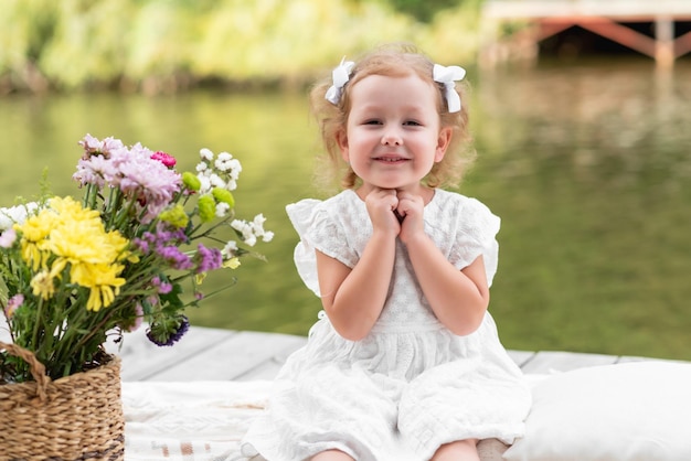 Kleines Mädchen in einem weißen Kleid sitzt auf einer Brücke am Fluss. Wir hatten ein Picknick am Wasser. Lächelnd und in die Kamera schauend