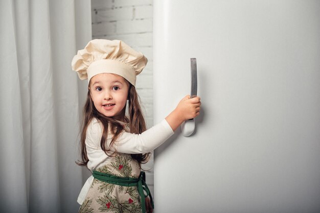 Kleines Mädchen in einer Kochmütze schaut in den Kühlschrank