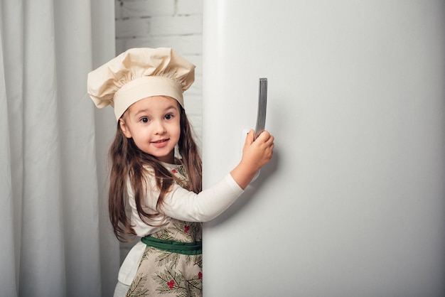 Kleines Mädchen in einer Kochmütze schaut in den Kühlschrank