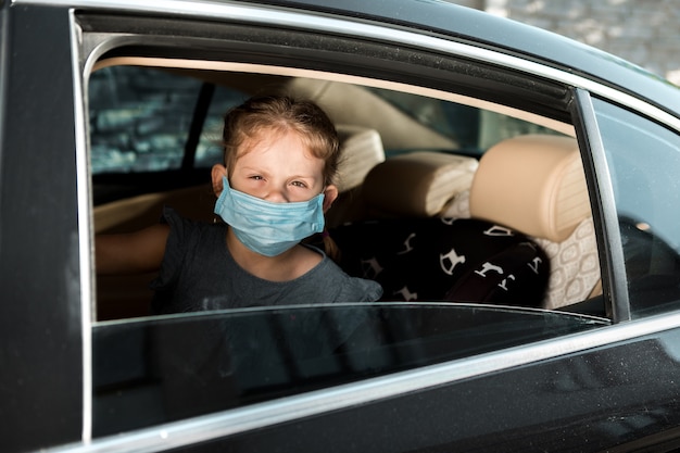 Kleines Mädchen in einer medizinischen Maske sitzt auf einem Kindersitz im Auto.