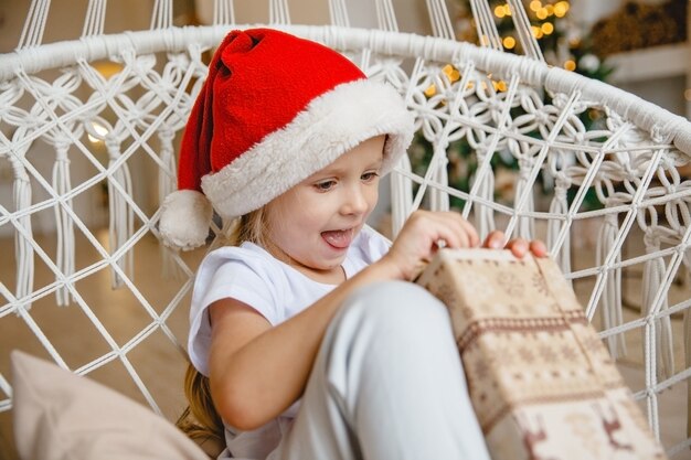 Kleines Mädchen in einer Weihnachtsmütze sitzt in einem Korbstuhl vor dem Hintergrund eines Weihnachtsbaumes und packt ein Geschenk aus