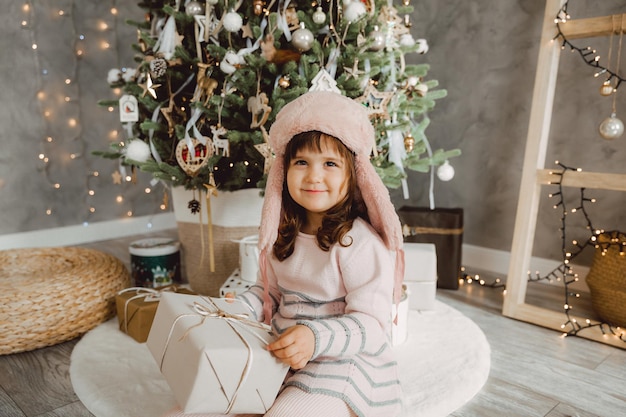 Kleines Mädchen in einer Winterpelzmütze sitzt mit Geschenken auf dem Boden neben dem Weihnachtsbaum.