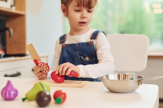 Kleines Mädchen in Freizeitkleidung sitzt am Tisch und hat Spaß beim Spielen mit Spielzeug in der Küche