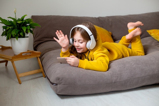 kleines Mädchen in gelber Kleidung liegt auf dem Bett und kommuniziert in sozialen Netzwerken über Smartphone