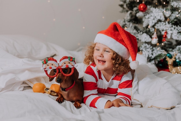 Kleines Mädchen in gestreiftem Pyjama und Weihnachtsmütze und ein Hund in lustigen Gläsern mit dem Weihnachtsmann