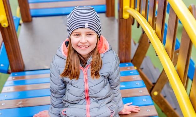 Kleines Mädchen in Mantel und Hut, die auf buntem hölzernen Spielplatz in der Herbstzeit sitzen