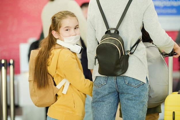 Kleines Mädchen in Maske mit Rucksack, das in die Kamera schaut, während es in einer Warteschlange am Flughafen steht