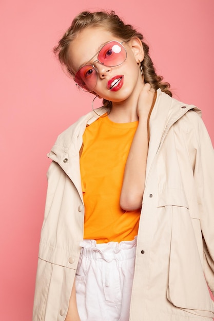 Kleines Mädchen in rosa Brille. Stilvolles Funtime-Konzept im trendigen Modestil. Vertikales Porträt in voller Länge in voller Länge des lachenden süßen süßen entzückenden reizenden reizenden Mädchens lokalisierter rosa Hintergrund