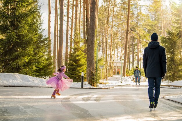 Kleines Mädchen in rosa Pullover und vollem Rock reitet am sonnigen Wintertag auf einer Eisbahn im Freien im Park