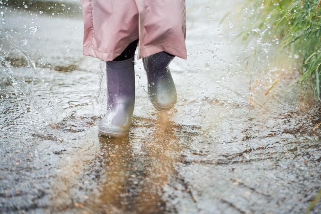 Kleines Mädchen in rosafarbenem wasserdichtem Regenmantel lila Gummistiefel lustige Sprünge durch Pfützen auf der Straße bei regnerischem Wetter Frühling Herbst Kinderspaß nach Regen Freizeitaktivitäten im Freien