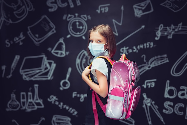 kleines Mädchen in Schuluniform mit medizinischer Maske mit Rucksack auf Tafelhintergrund