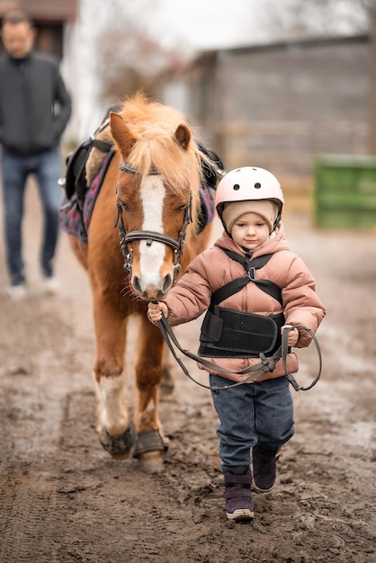 Kleines Mädchen in Schutzjacke und Helm mit ihrem braunen Pony vor der Reitstunde