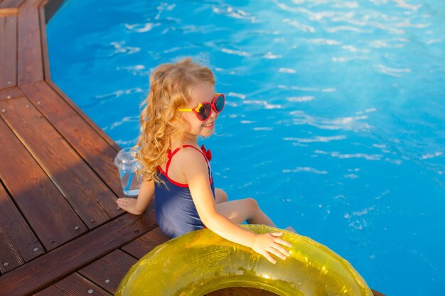 Kleines Mädchen in Sonnenbrille und Badeanzug, im Sommer am Pool sitzend