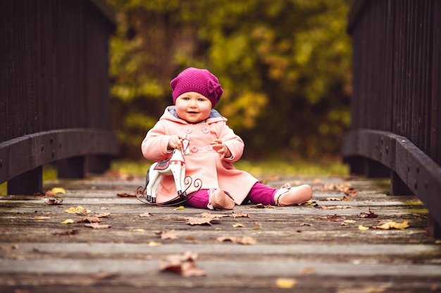 Kleines Mädchen in Strickmütze und Jacke sitzt auf der Holzbrücke mit viel Herbstlaub im Park