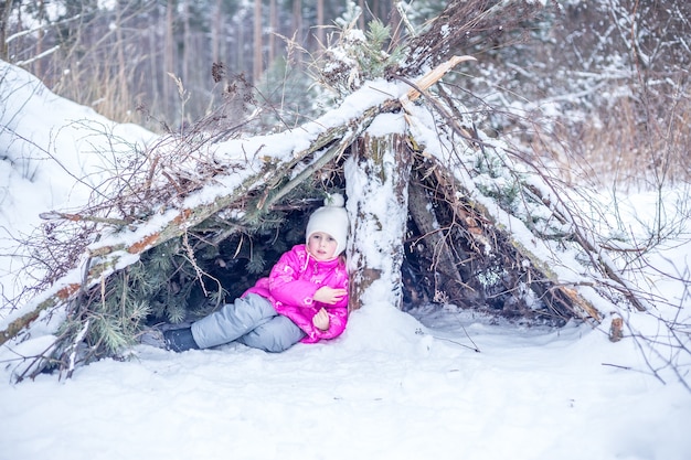 Kleines Mädchen in warmer Kleidung spielt in einer Hütte aus Nadelzweigen im Winterwald und verbringt im Winter Zeit im Freien